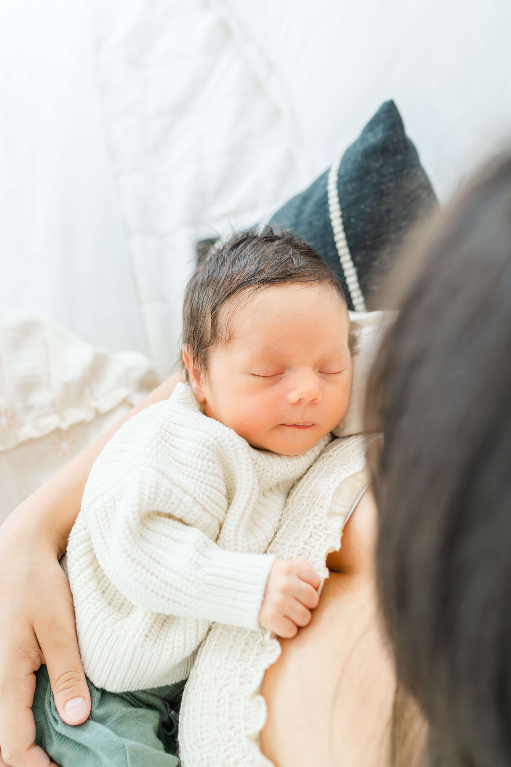 A newborn baby in a white knit sweater sleeps against mom's chest on a bed after finding nannies in austin