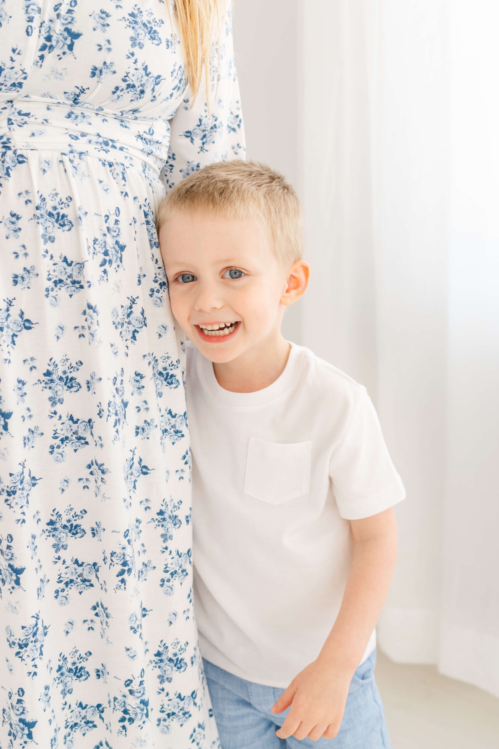 A smiling toddler boy in a white shirt hugs mom's leg in a blue floral dress while standing in a studio after meeting a pediatric dentists in austin