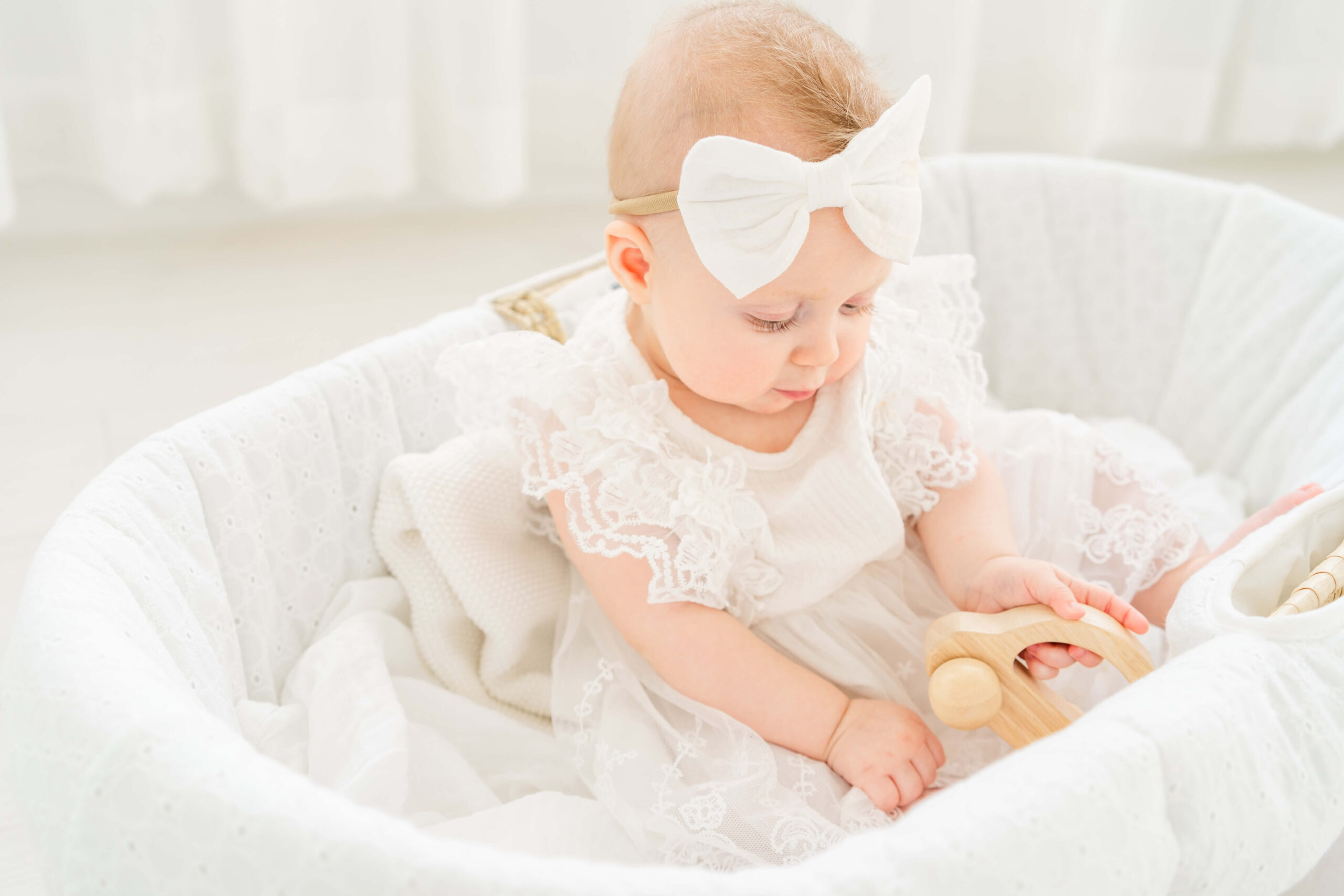 A baby girl in a white dress plays with a wooden car in a small crib after visiting pediatric dentists in austin