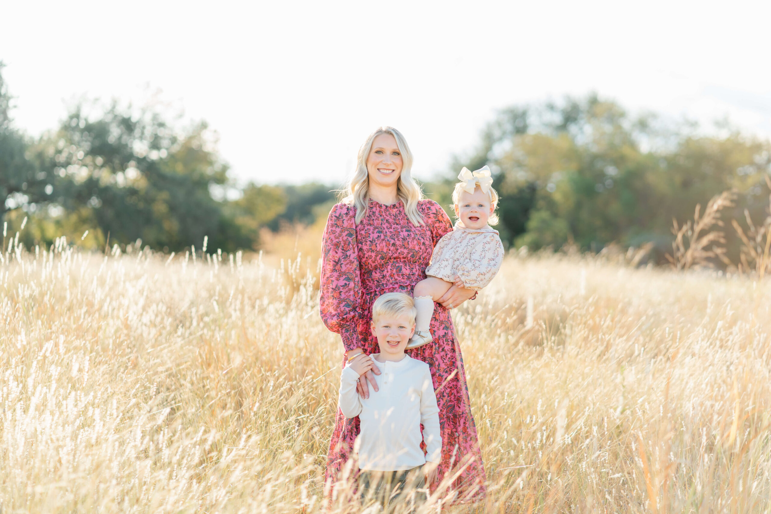 A smiling mother in a pink floral print dress stands with her toddler son and holding her younger daughter on her hip
