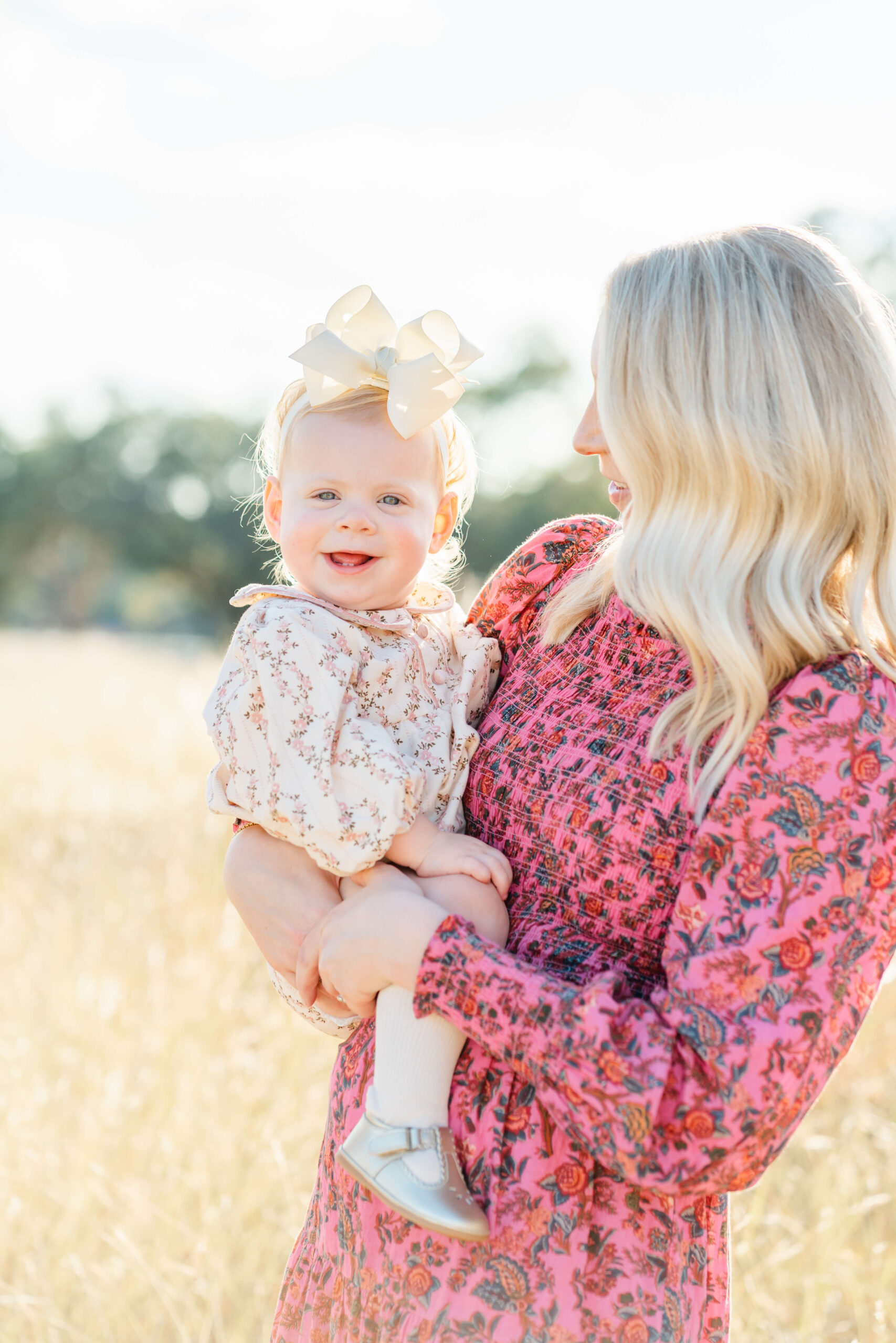 A happy toddler baby girl smiles big sitting in mom's arms in floral print dresses in a field of tall grass after meeting pediatricians in austin
