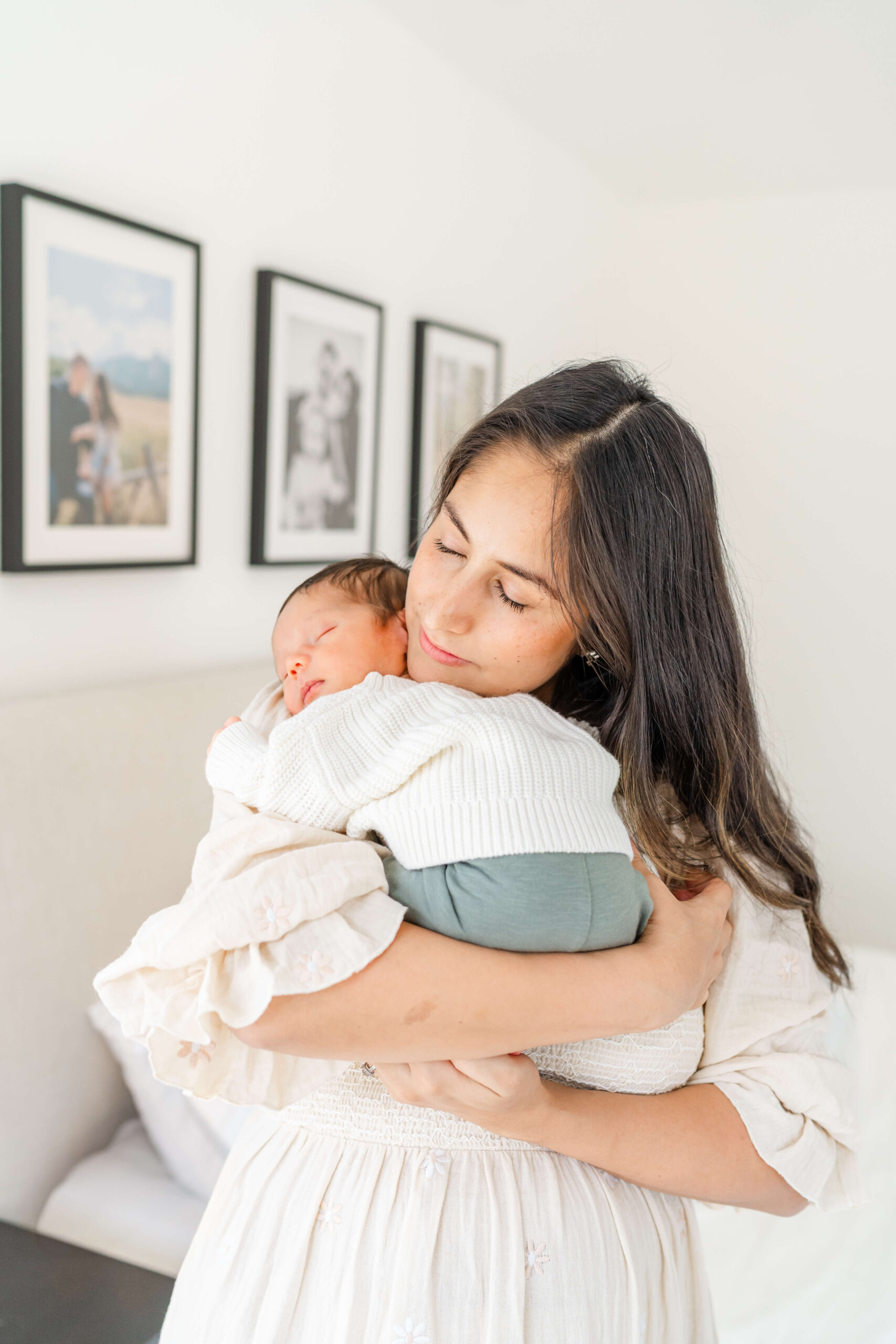 A smiling new mom in a white dress cradles her sleeping newborn under her chin while standing in a bedroom after meeting postpartum doulas in austin