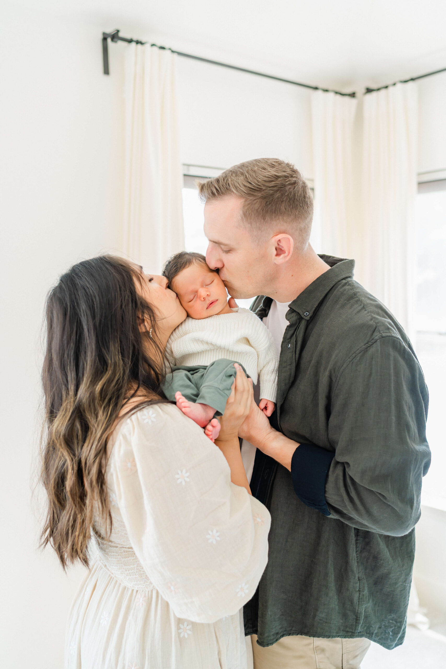 a mom and dad in white and green kiss the cheeks of their sleeping newborn between them after meeting postpartum doulas in austin