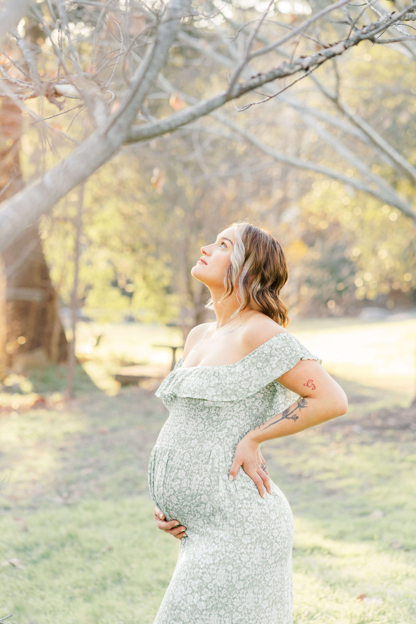 A mother to be stands in a park at sunset with a hand under her bump and looking up to a tree