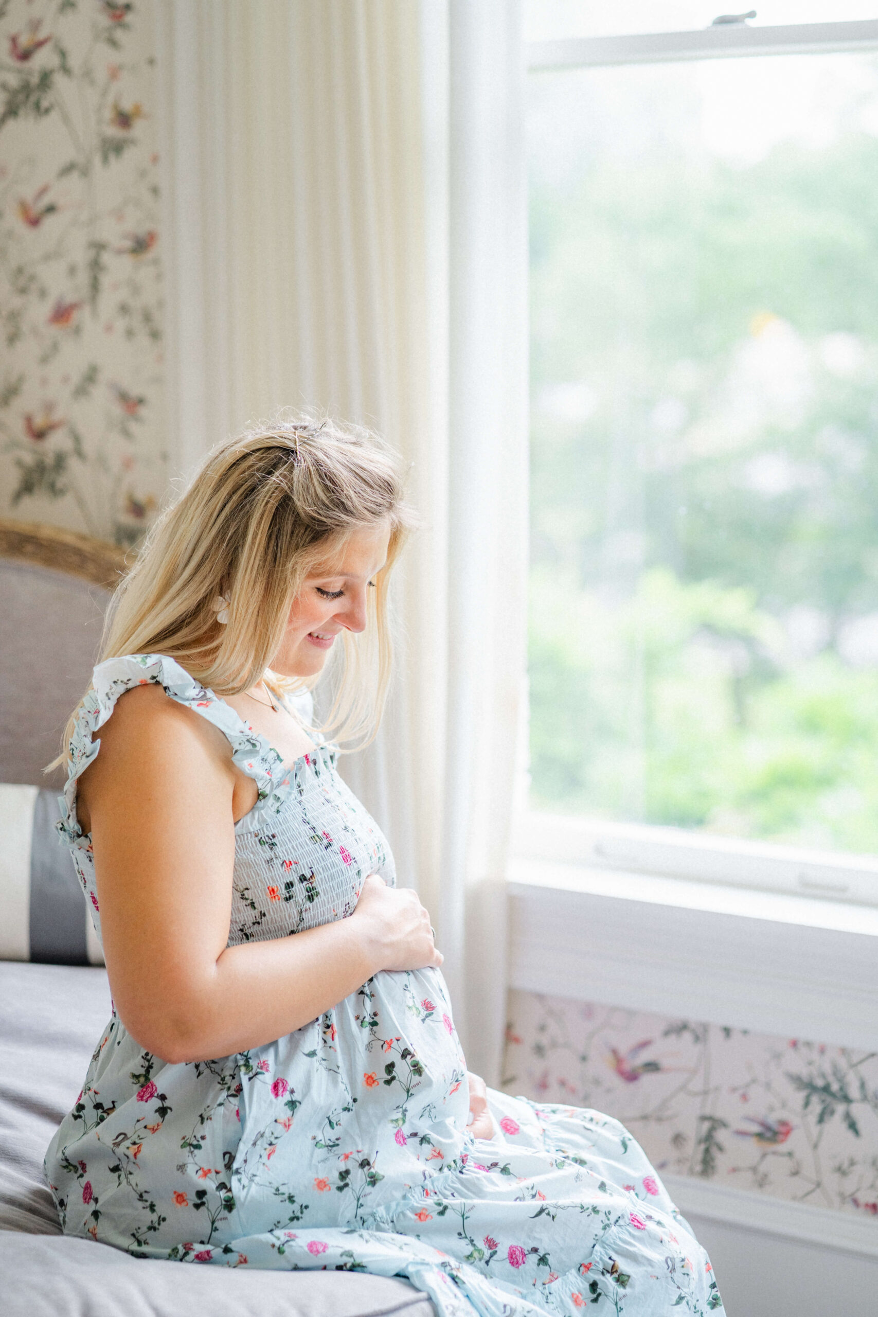 A happy pregnant woman sits on a bed under a window smiling down to her bump in a blue maternity dress