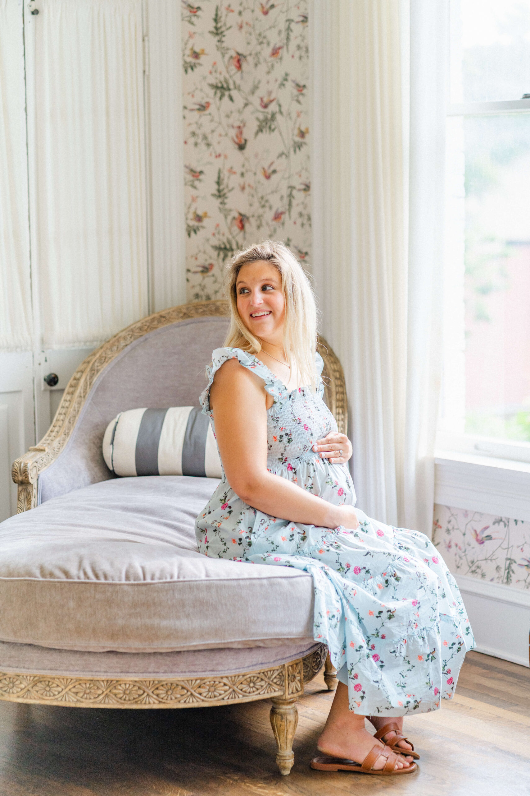 A pregnant woman smiles over her shoulder while sitting on a vintage lounger under a window in a blue dress after exploring doulas in austin