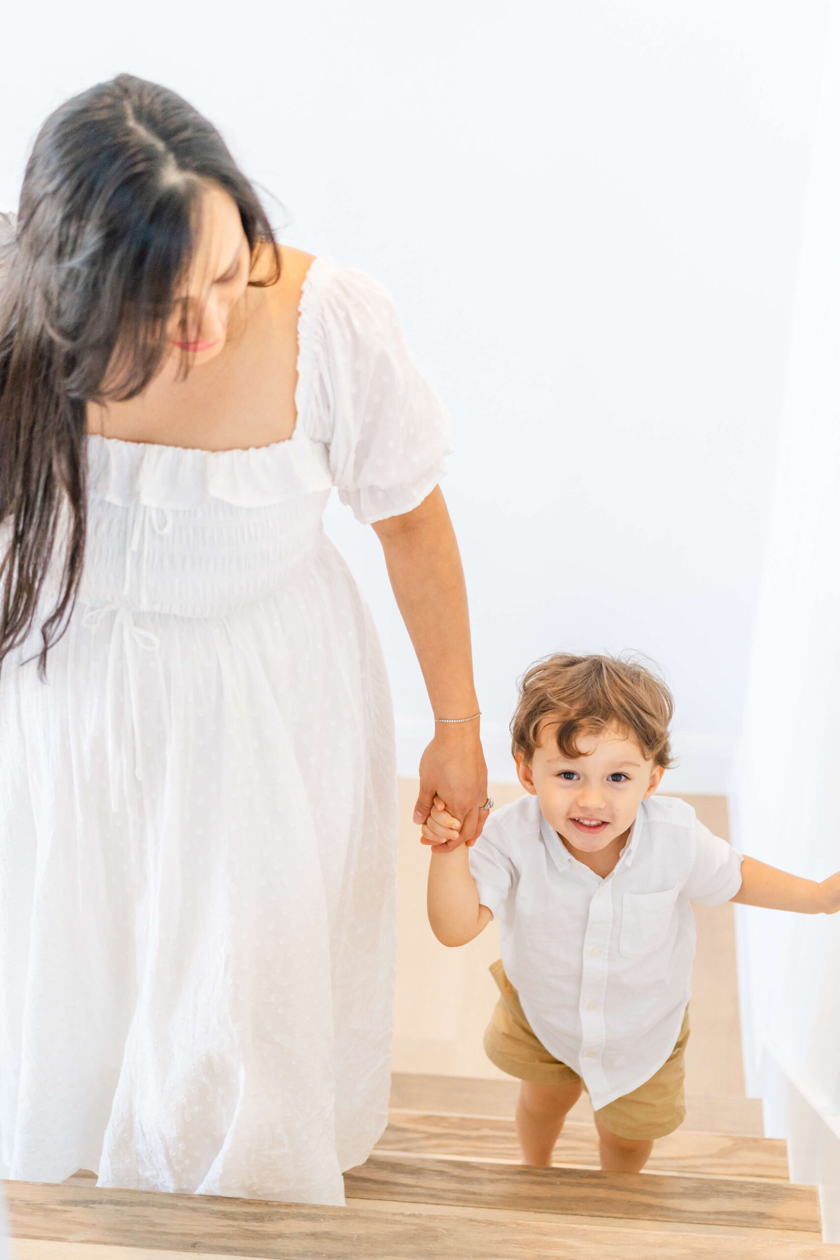 A mom in white leads her toddler son up wooden stairs by the hand with smiles after using obgyns in austin