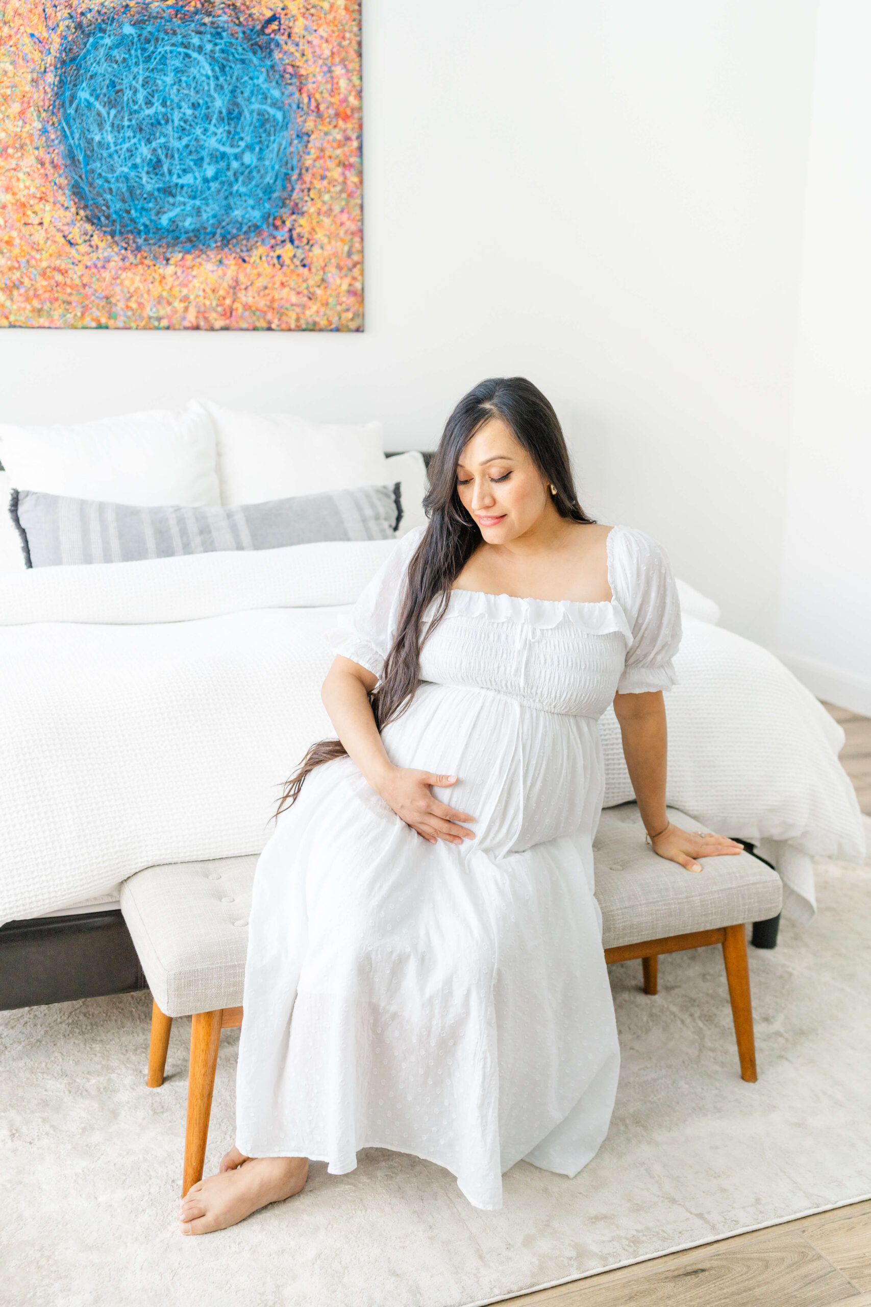A pregnant woman in a white maternity gown sits on a bench with a hand under her bump in a bedroom