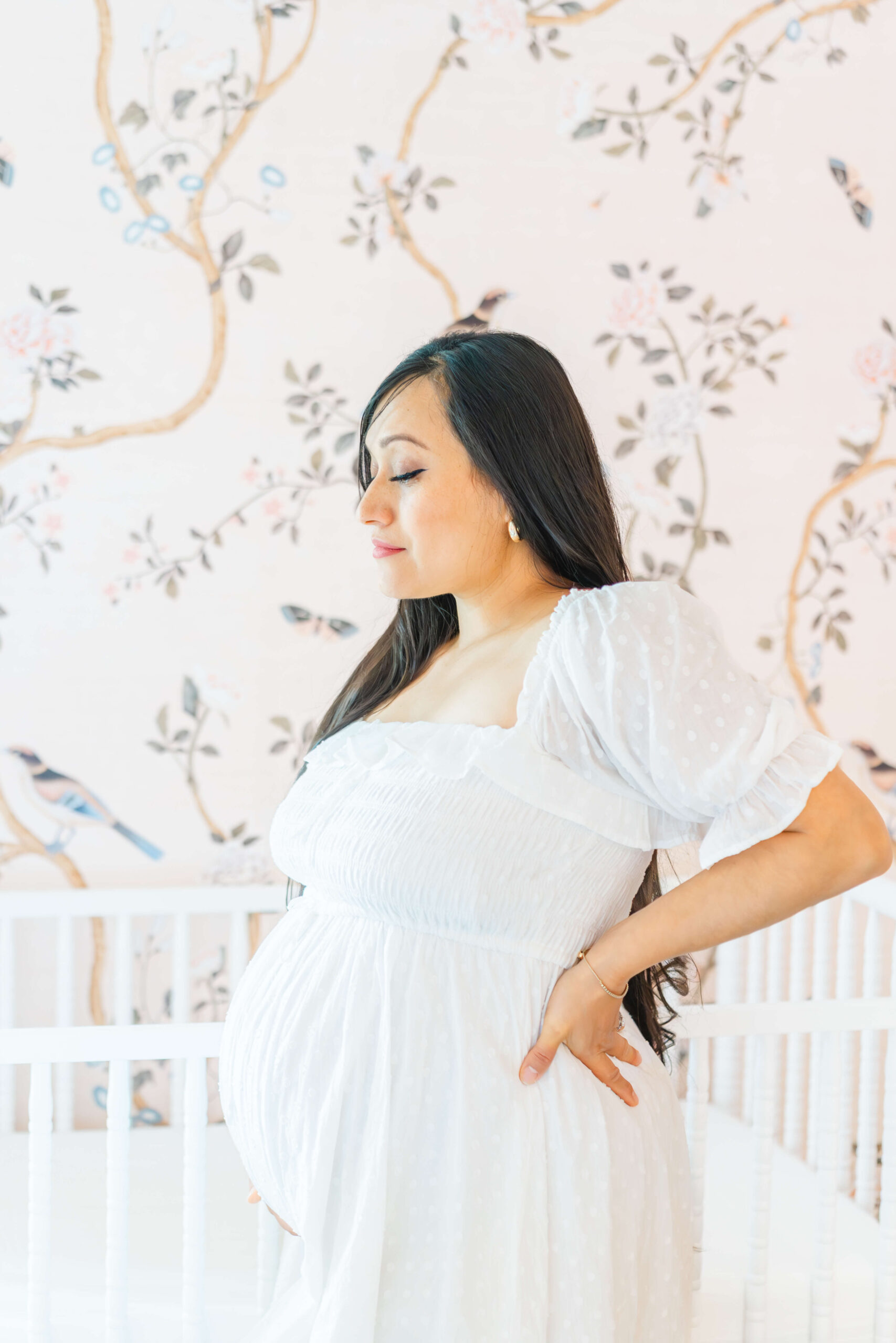 A pregnant woman in a white maternity gown stands in a nursery with a hand on her hip after visiting obgyns in austin