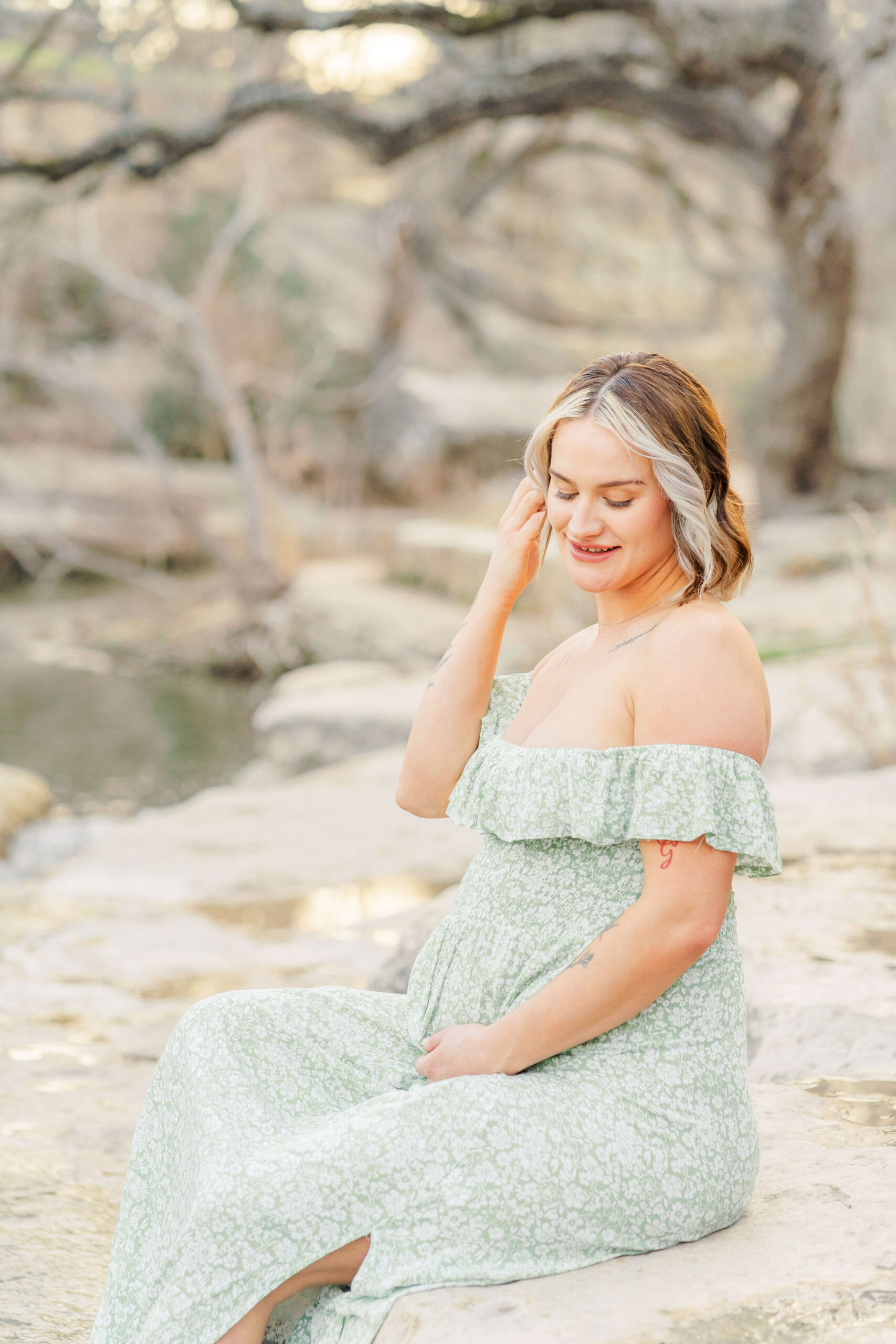 A happy pregnant woman sits on the edge of a creek pulling her hair back in a green maternity gown