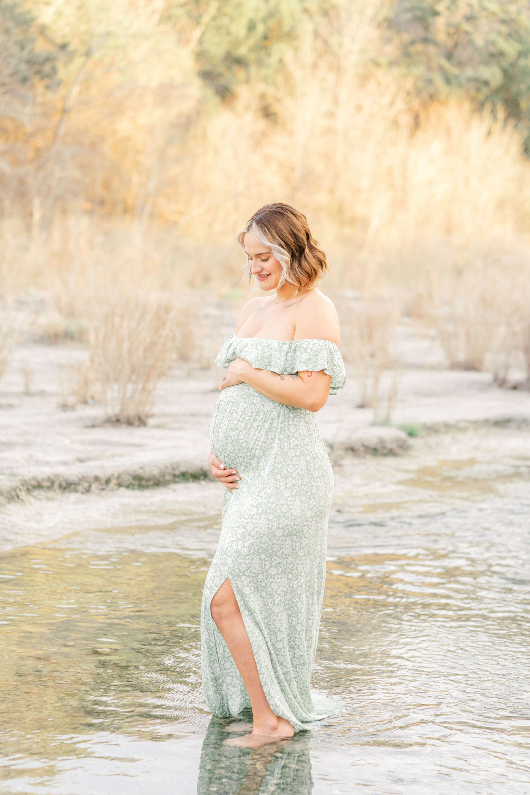 A mother to be walks in a shallow creek in a green maternity gown smiling down to her hands on her bump after visiting prenatal chiropractors in austin