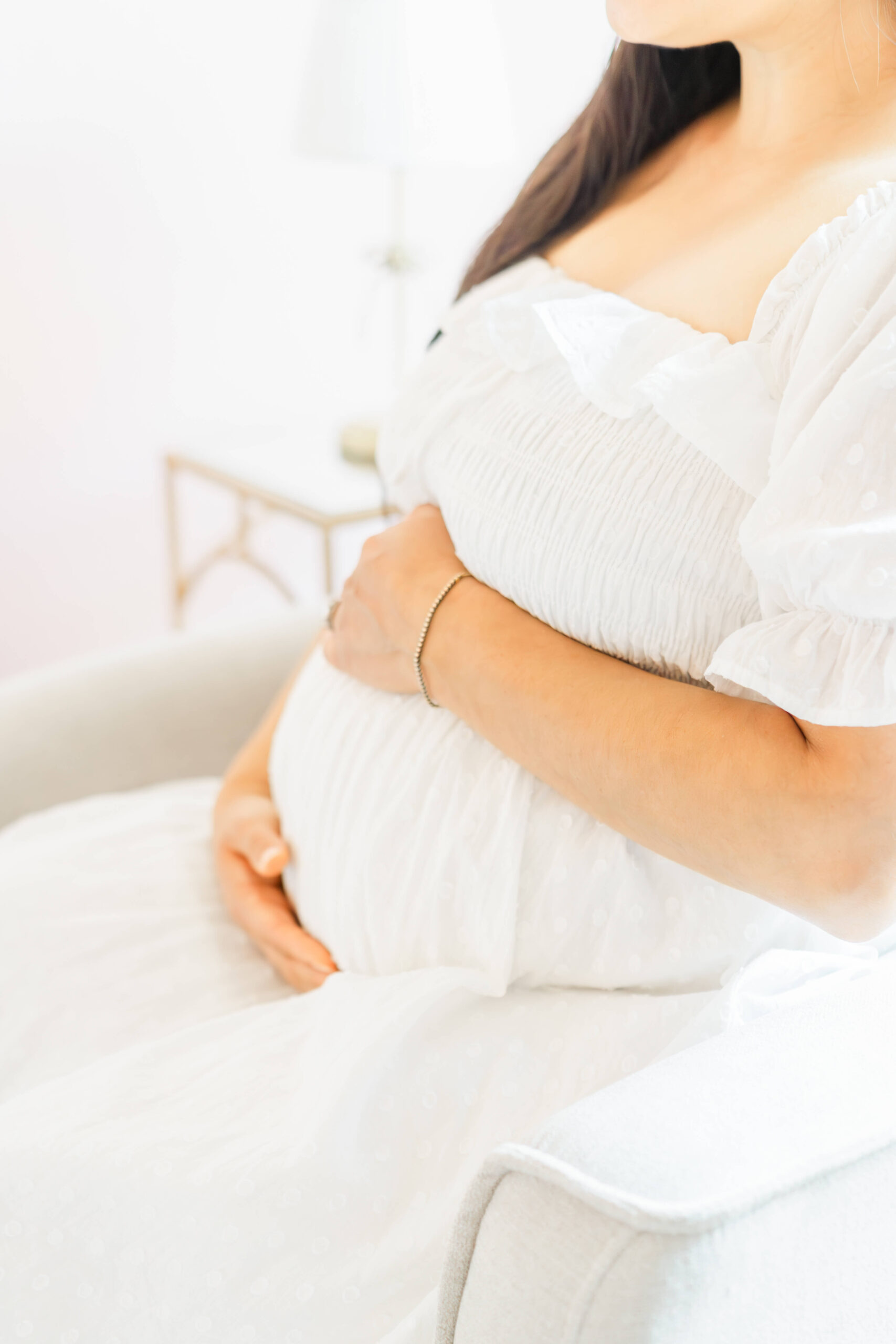 Details of a pregnant woman sitting in a chair holding her bump in a white maternity dress