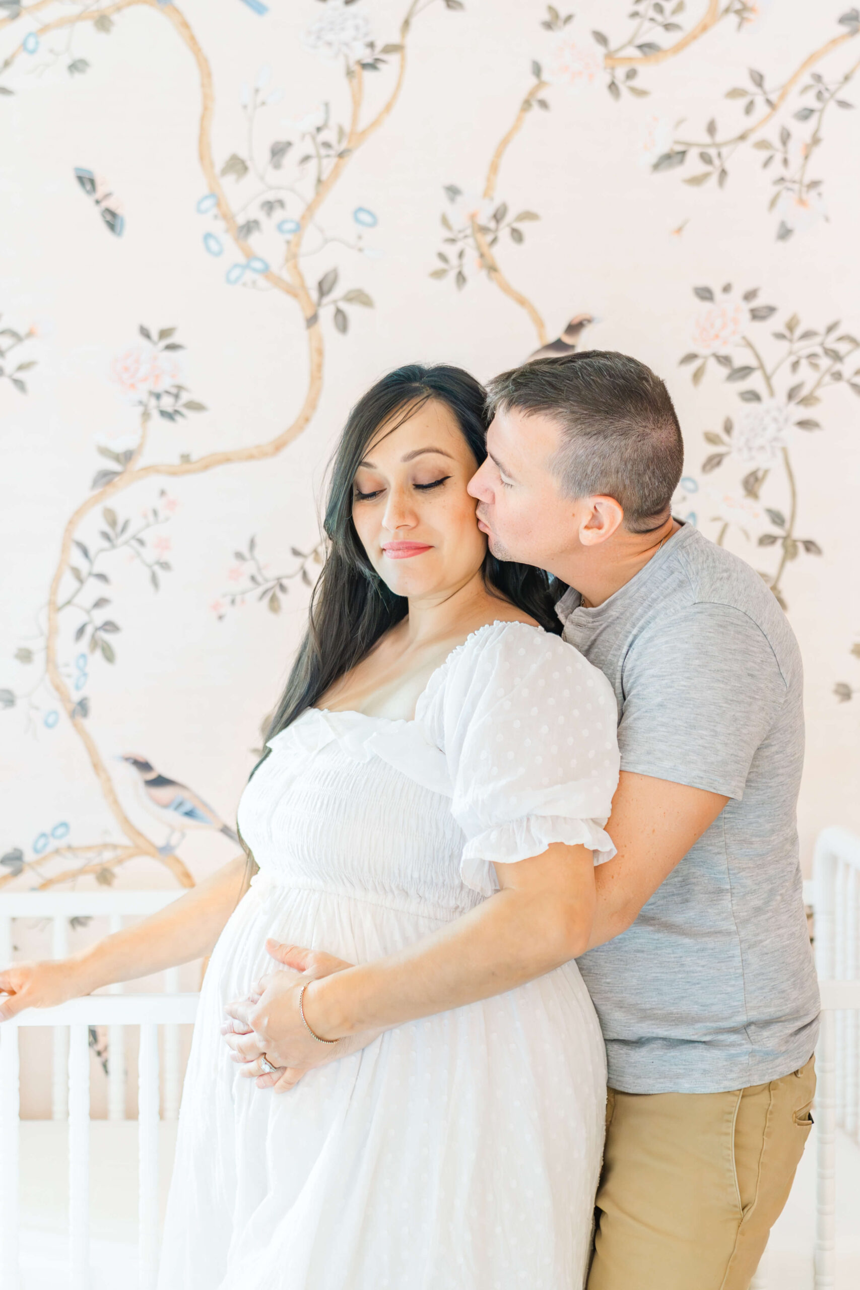 A man kisses the cheek of his pregnant wife in a white maternity gown while holding the bump and leaning on a crib in a nursery after a prenatal massage in austin