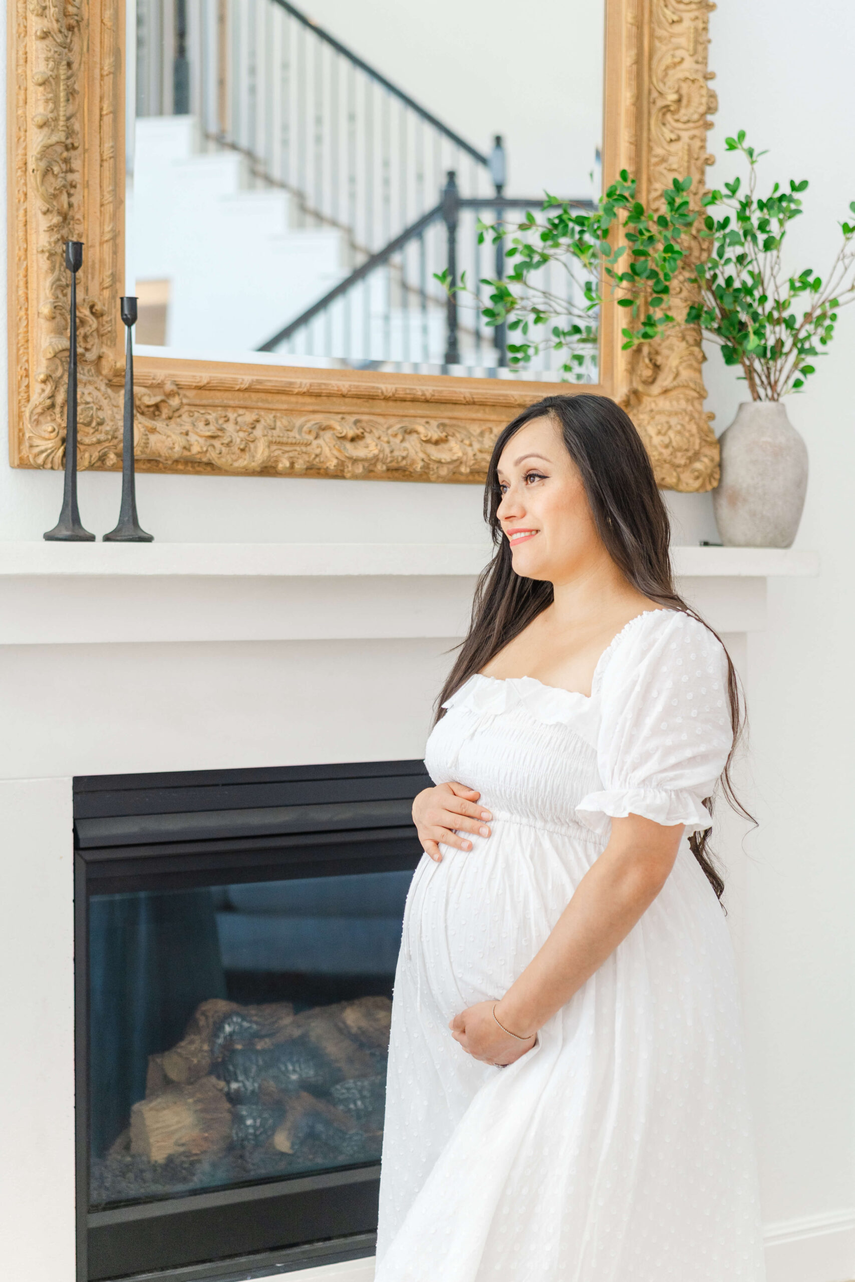 A smiling mother to be stands in front of the fireplace in a white maternity gown holding her bump