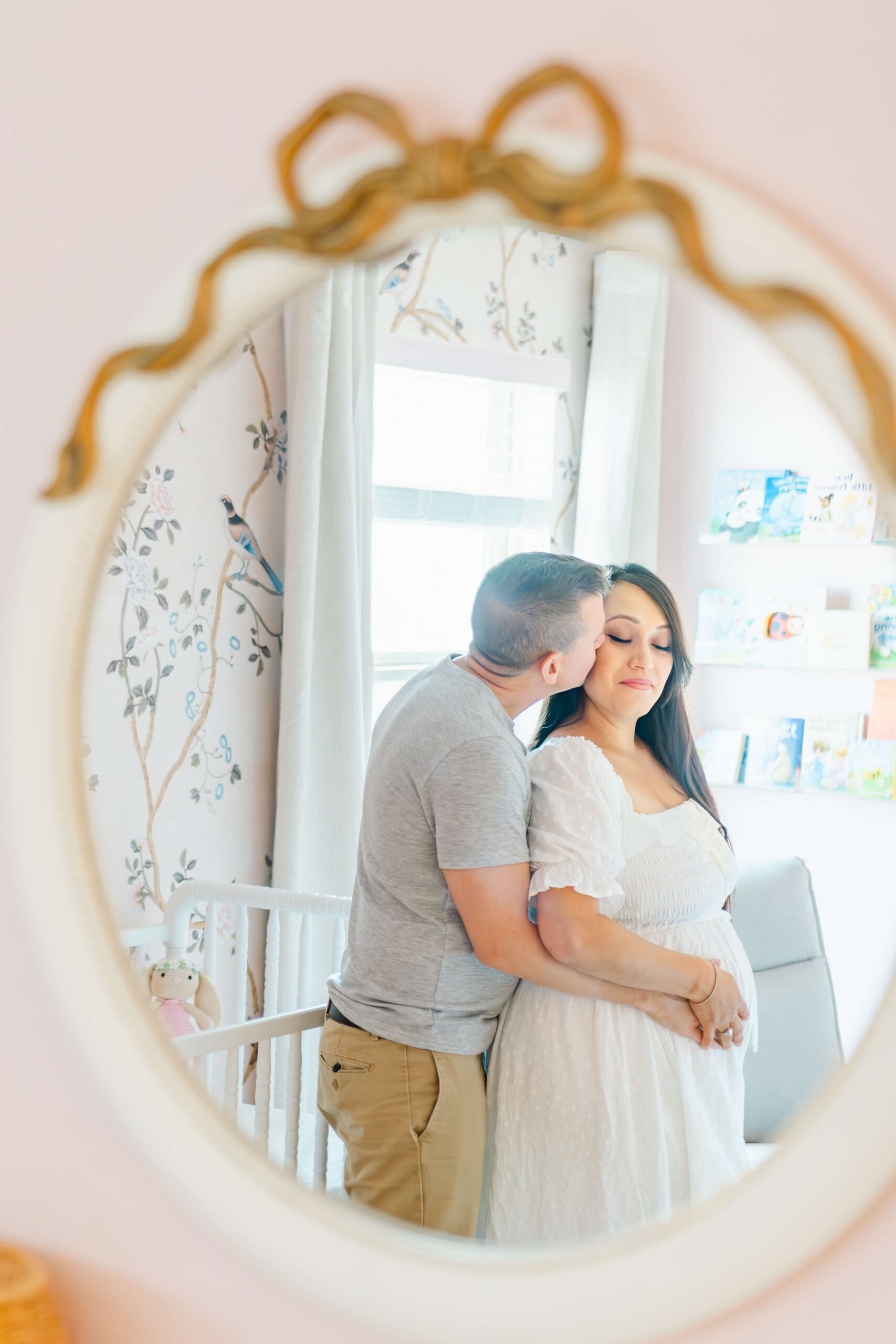 A happy mom to be is kissed on the cheek by her partner in a nursery in a white maternity dress seen through a mirror after some prenatal in yoga austin