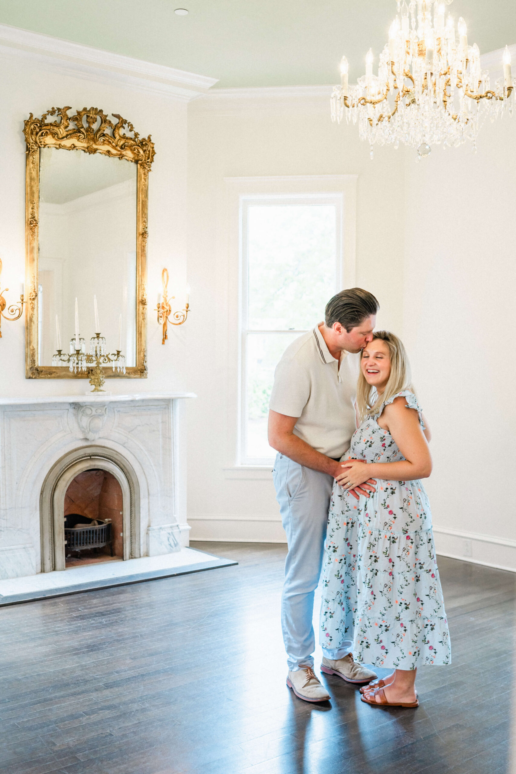 A happy expecting couple smile big and kiss the forehead in a luxurious room in a blue maternity dress and white shirt after visiting a Maternity Boutique in Austin