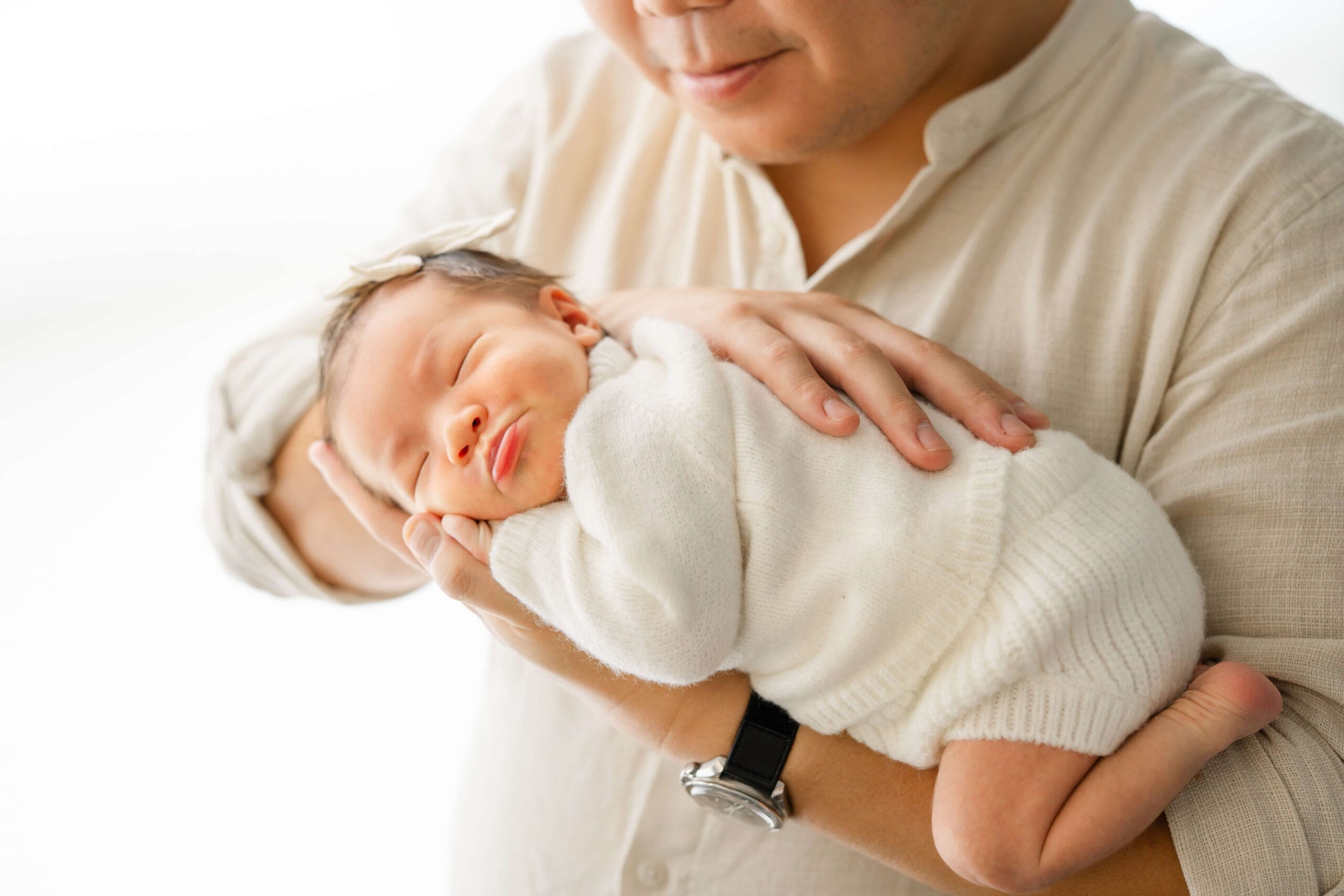 A new dad cradles his sleeping newborn on his arm in a white sweater