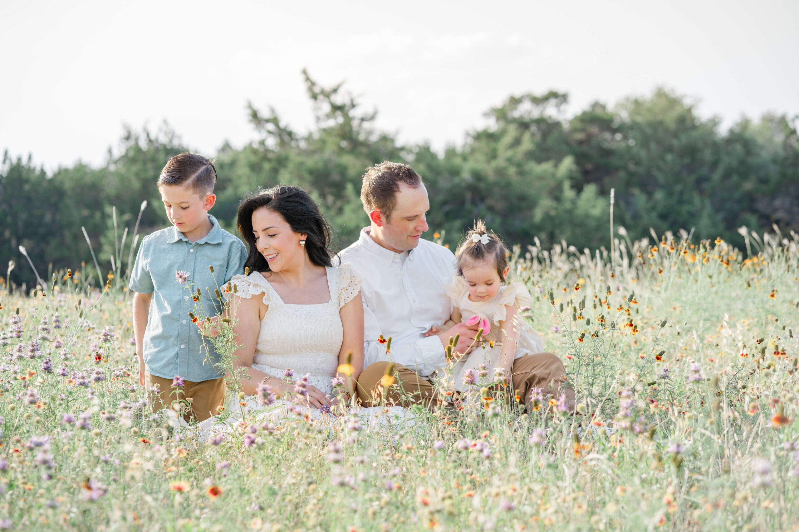 A happy mom and dad sit in a field of wildflowers picking them with their toddler son and daughter in their laps after visiting infant daycares in austin
