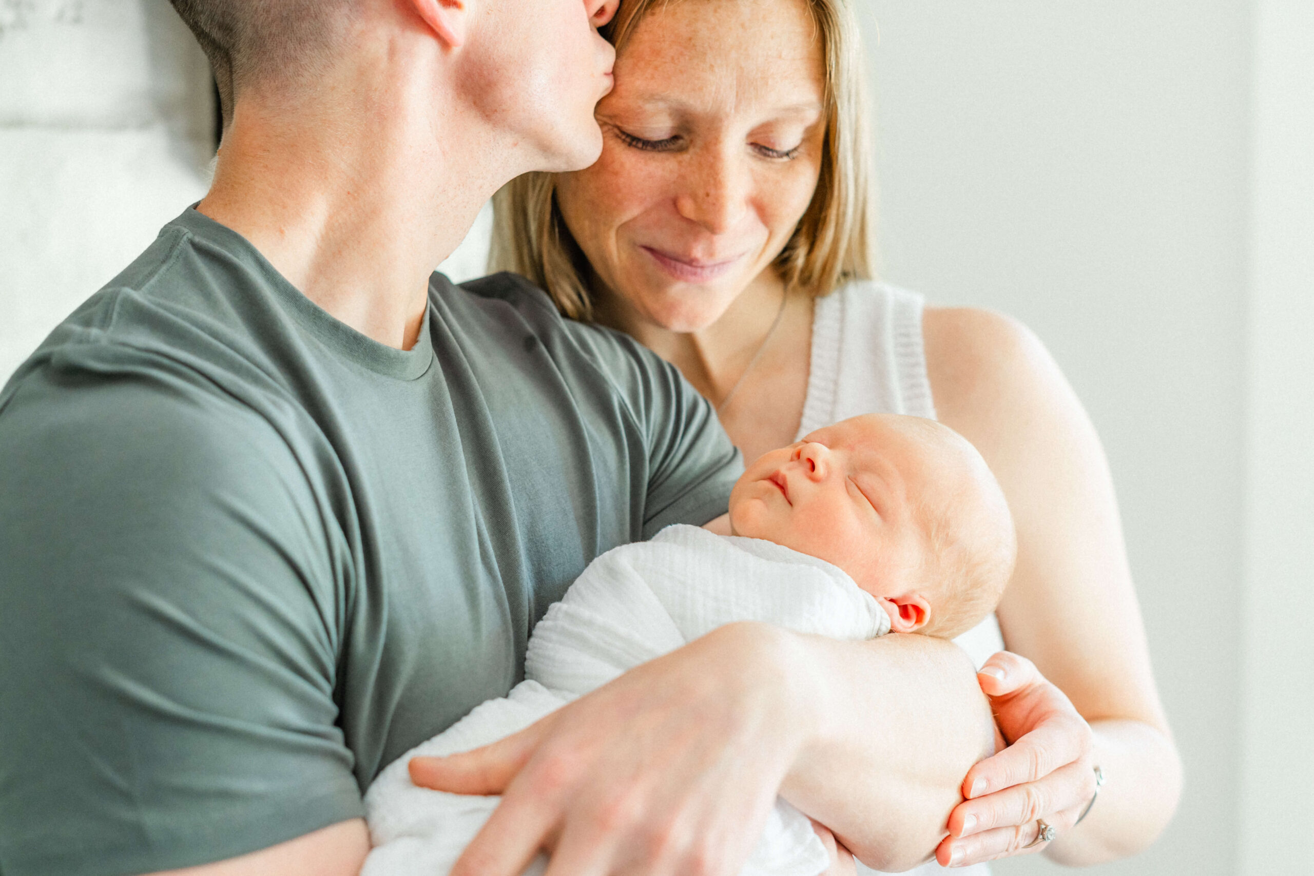 A mom and dad smile and kiss their sleeping newborn baby in dad's arms in a white swaddle
