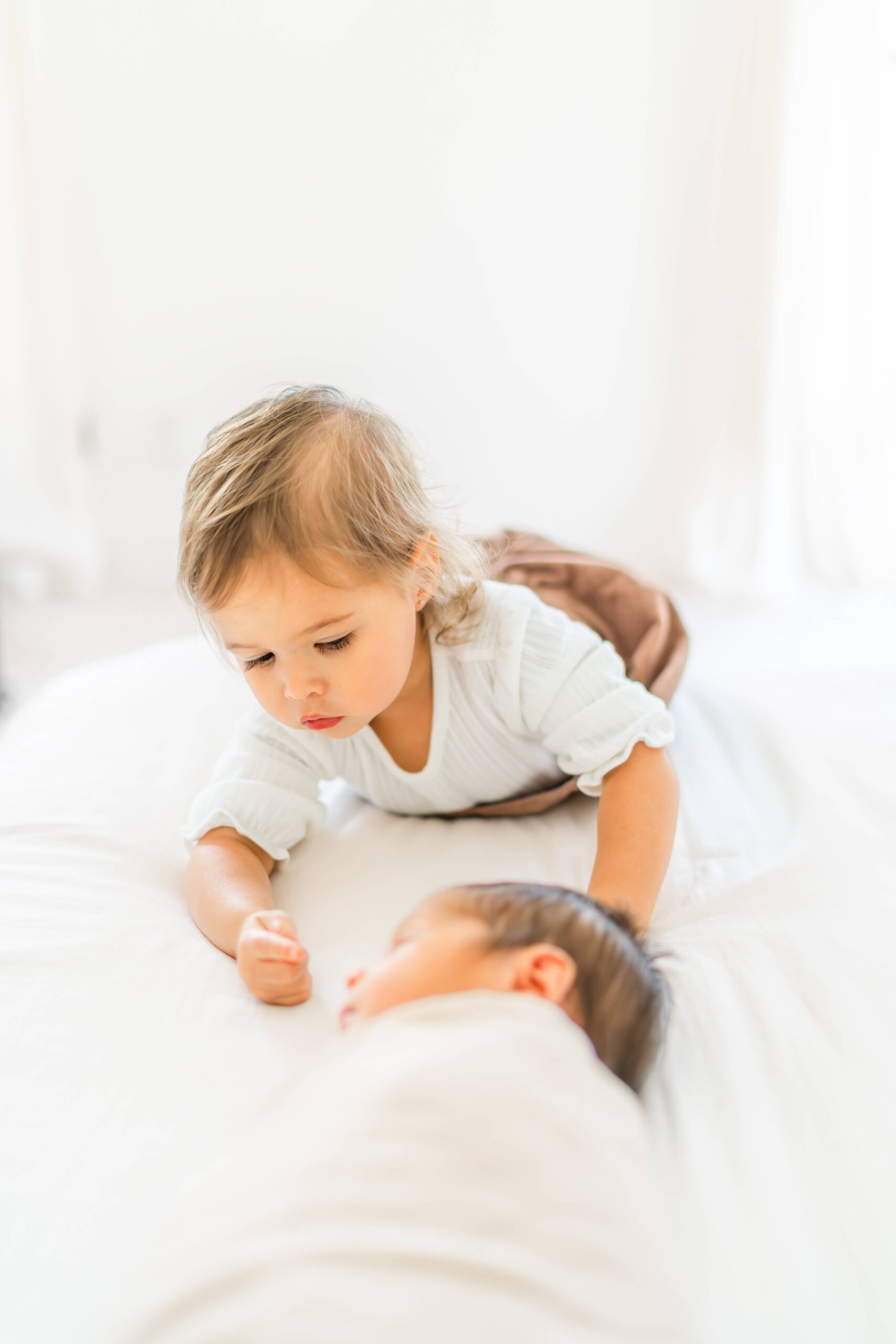 A toddler in a white shirt crawls on a bed to meet her newborn baby sibling in a white swaddle after visiting pediatric chiropractors in austin