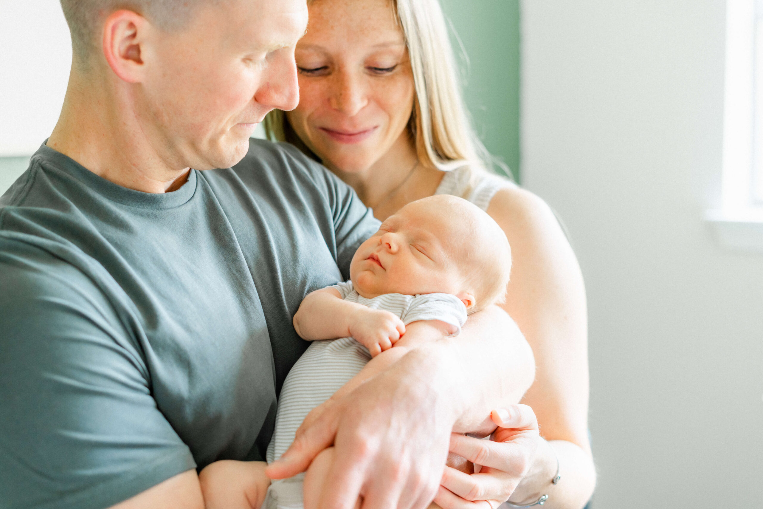 Smiling new mom and dad admire their sleeping newborn in a onesie in dad's arms while standing in a window
