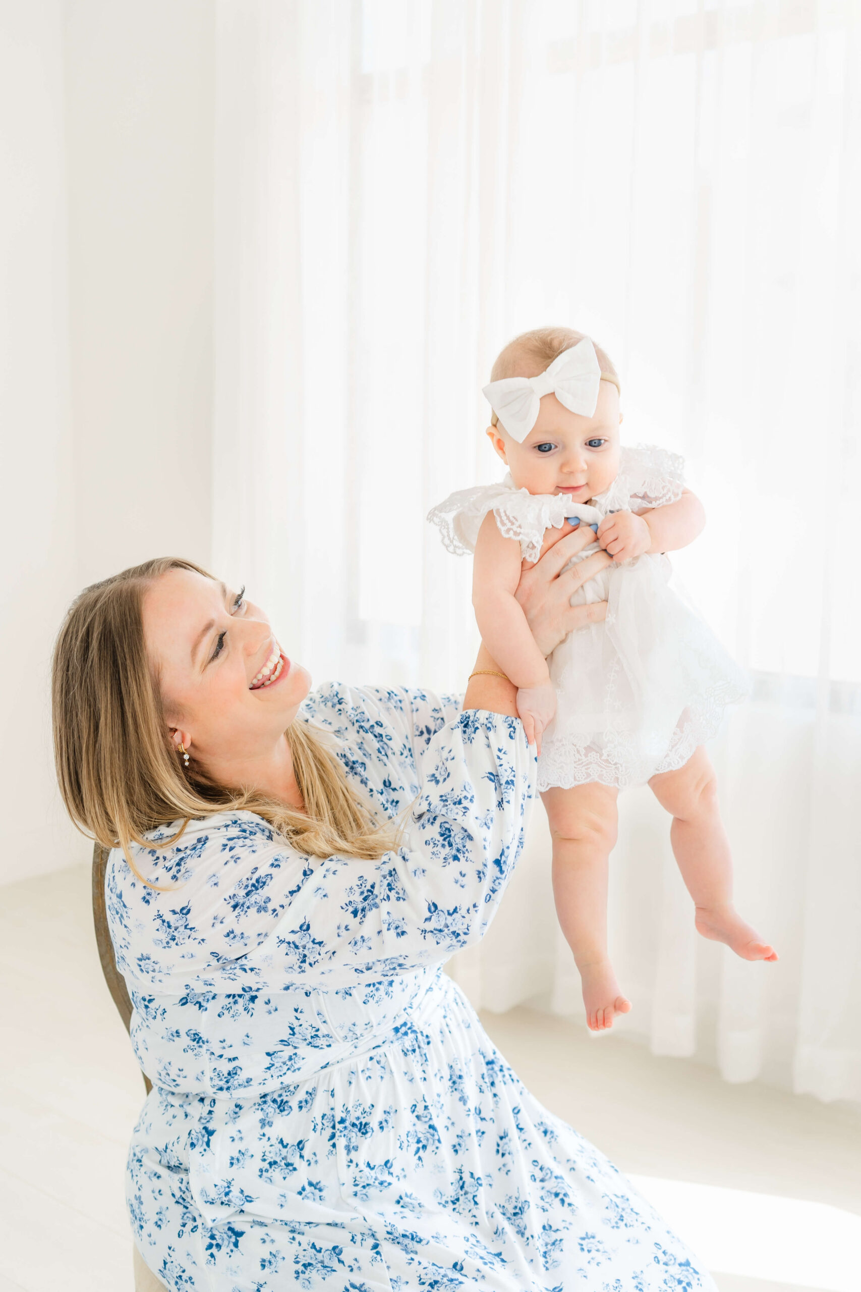 A happy new mom in a blue flower print gown lifts her baby in a white onesie in a studio after some baby music classes in austin