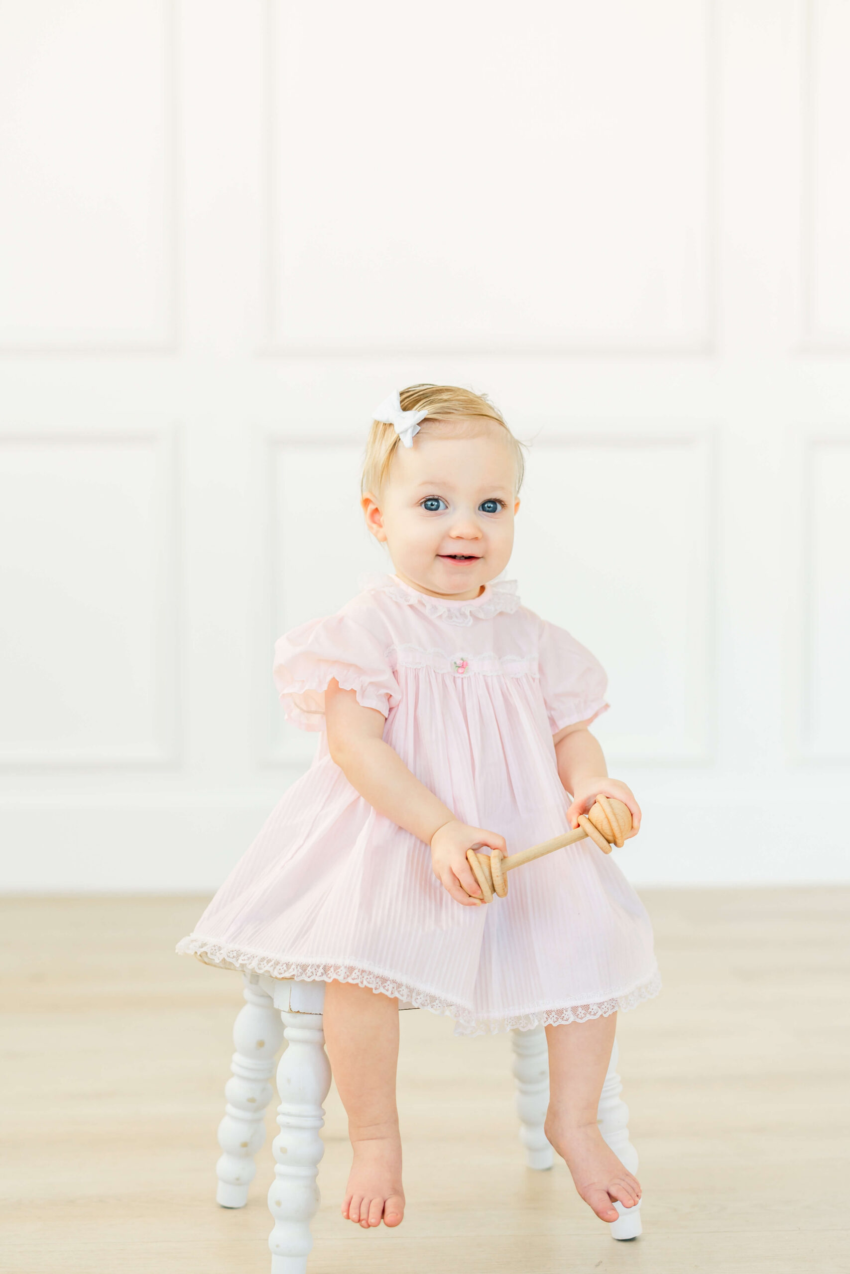 A happy baby girl sits on a wooden stool in a pink dress holding a wooden rattle before visiting indoor playgrounds in austin