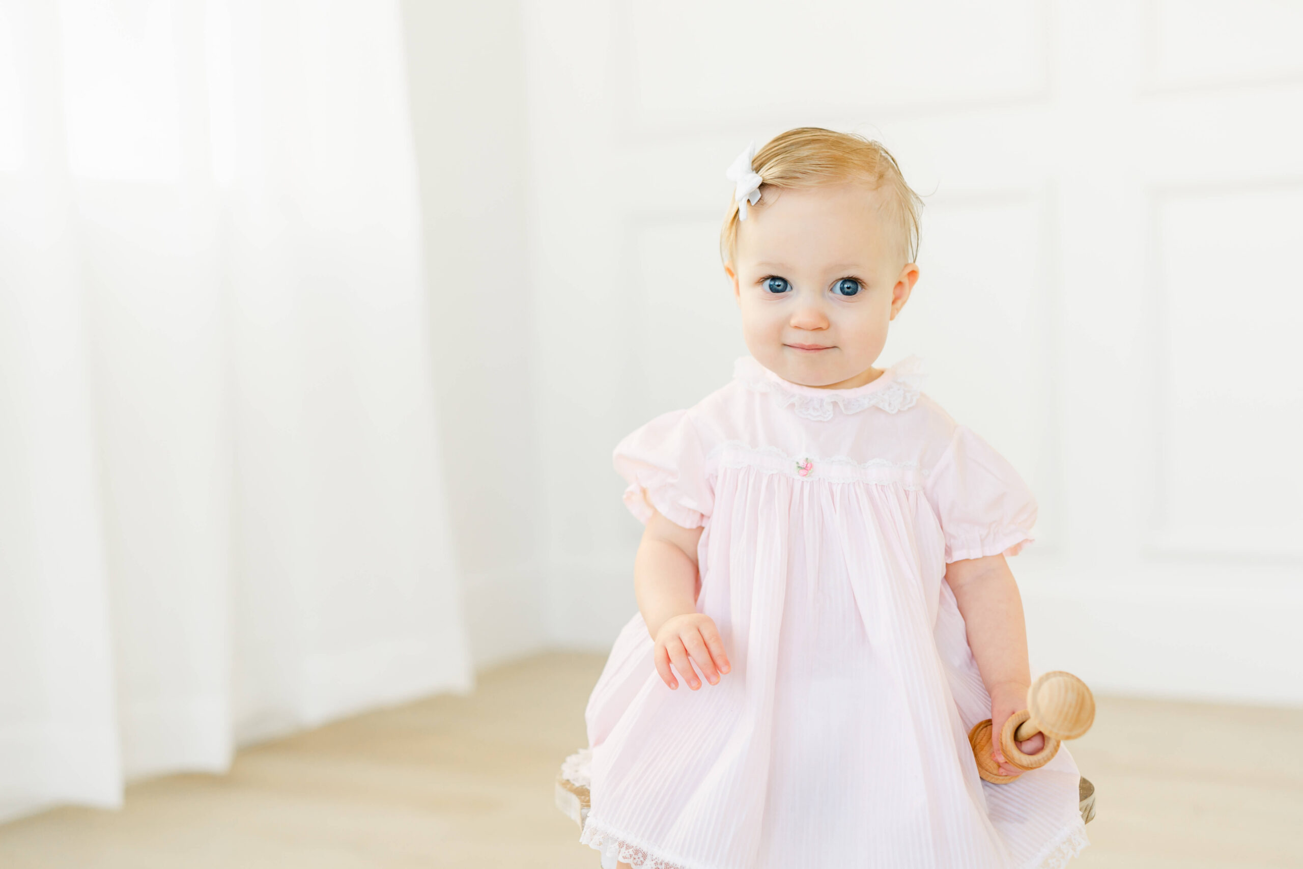 A happy toddler girl in a pink dress plays with a wooden rattle in a studio before visiting indoor playgrounds in austin