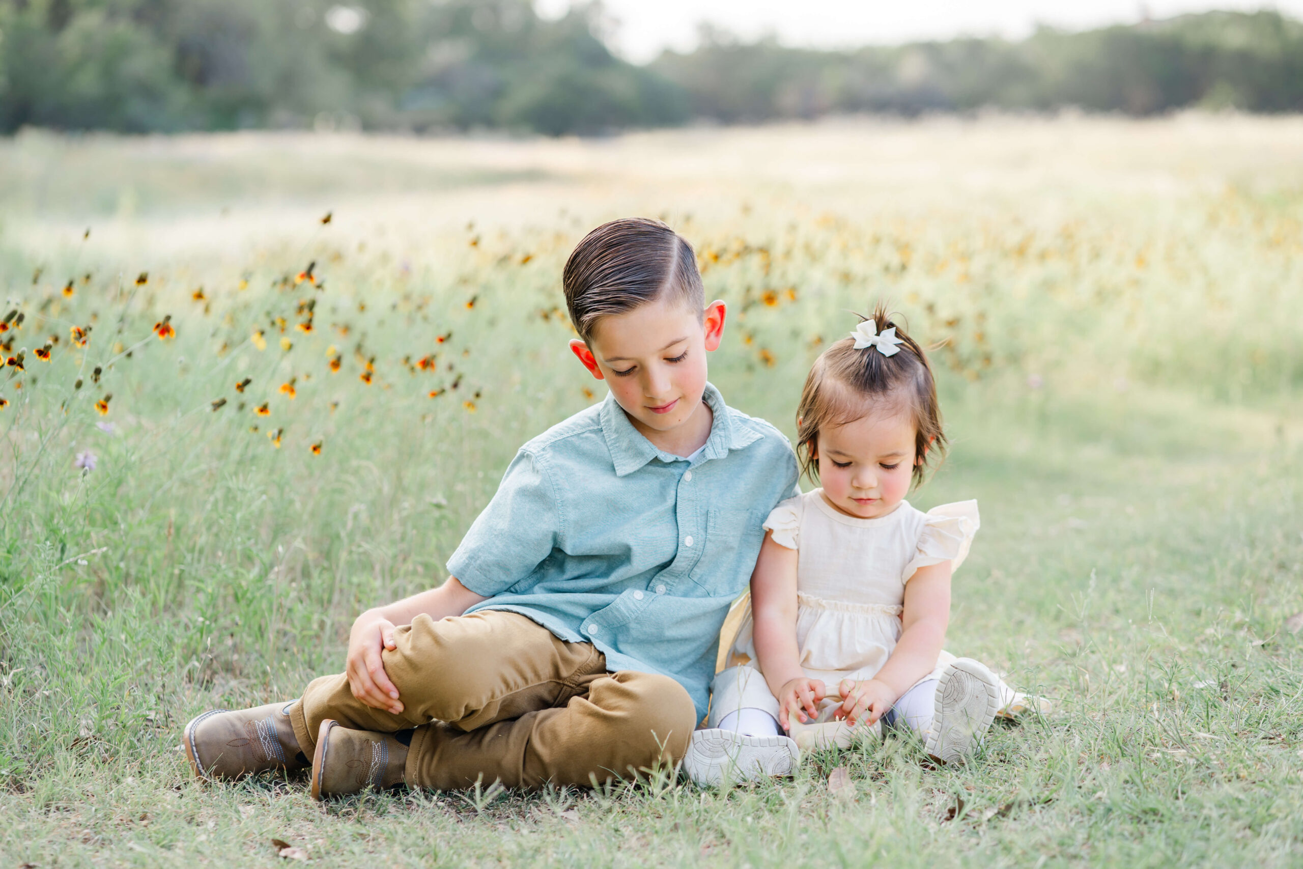 A young boy in a green button down sits in a path with her toddler sister playing with a toy surrounded by wildflowers
