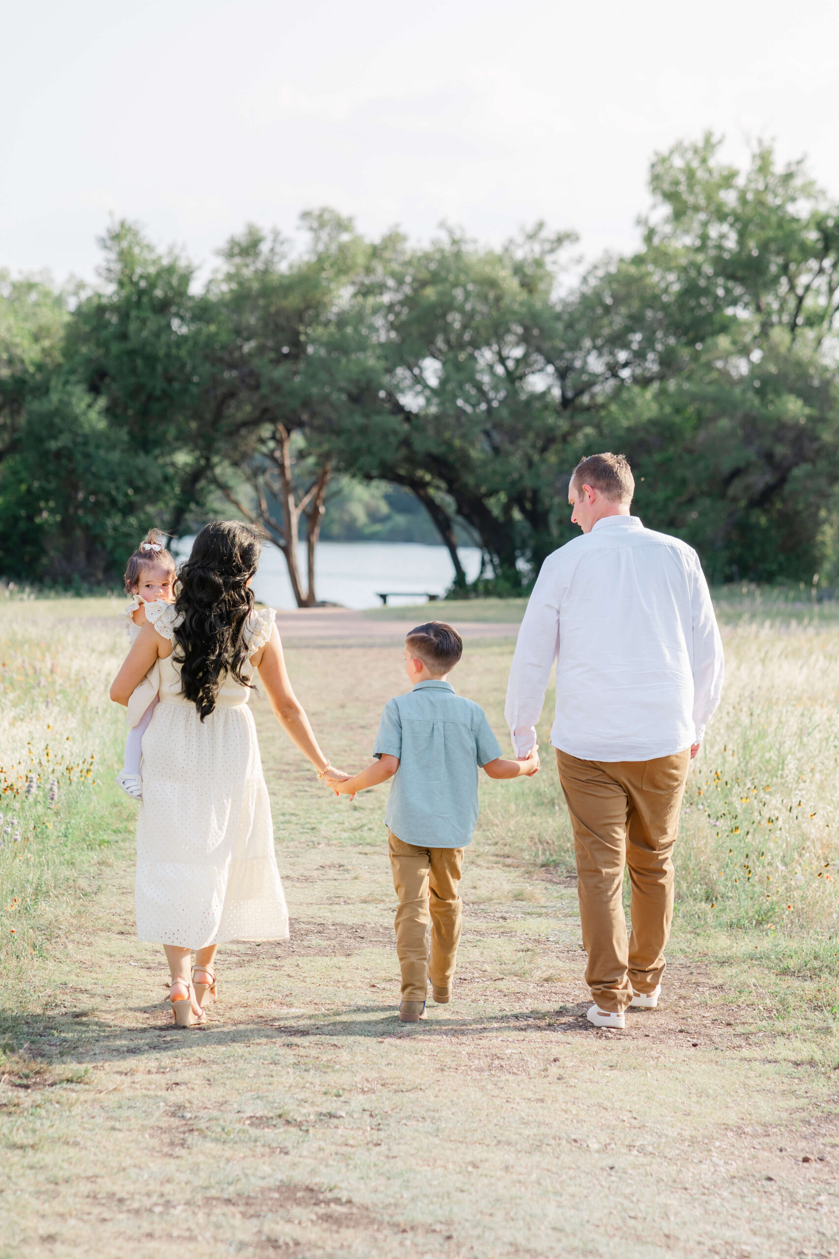 A mom and dad in white walk in a park to a lake holding hands with their young son and carrying a toddler girl after visiting montessori schools in austin