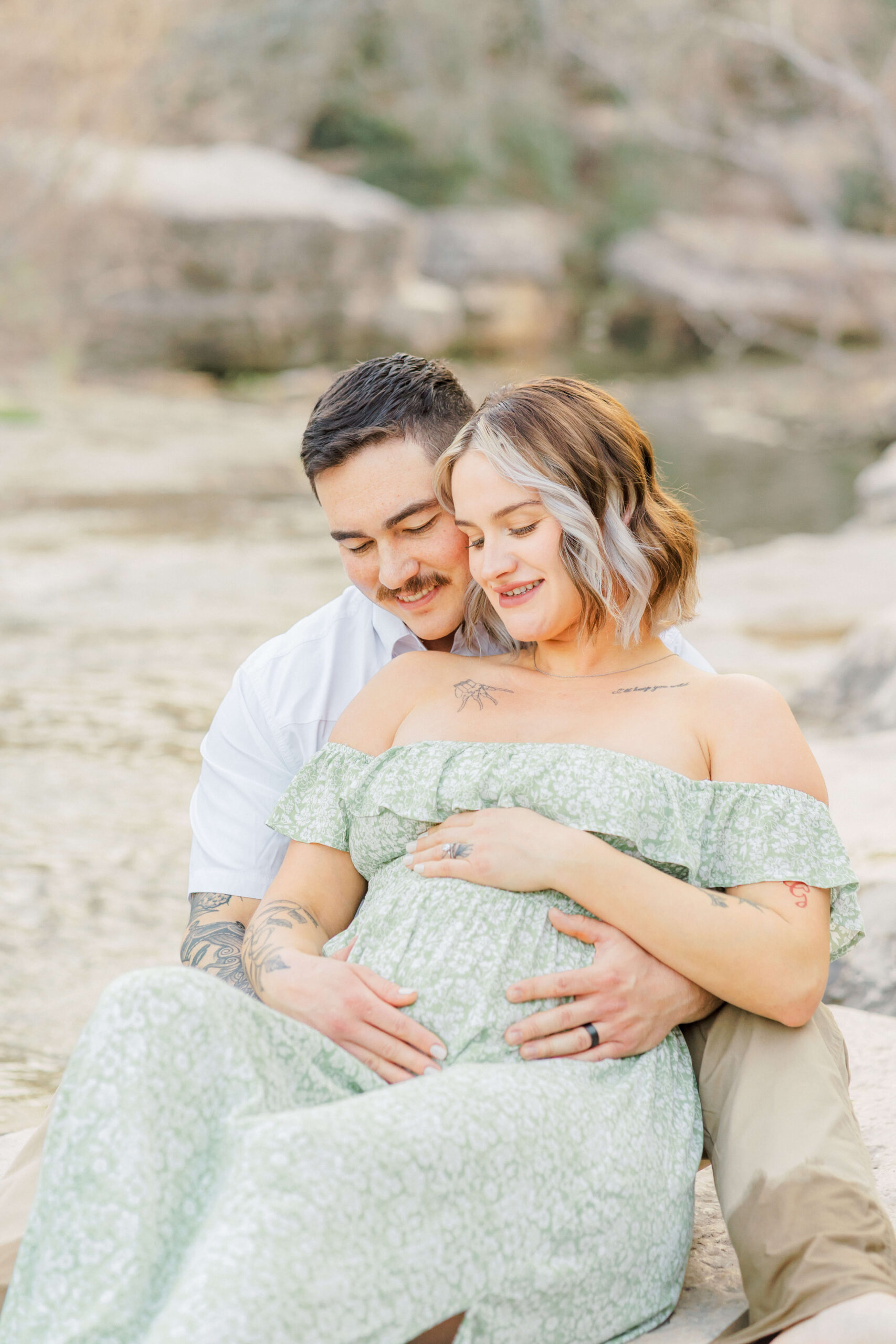 A happy expecting couple snuggles by a river in a green maternity gown smiling at the bump before needing pelvic floor therapy in austin