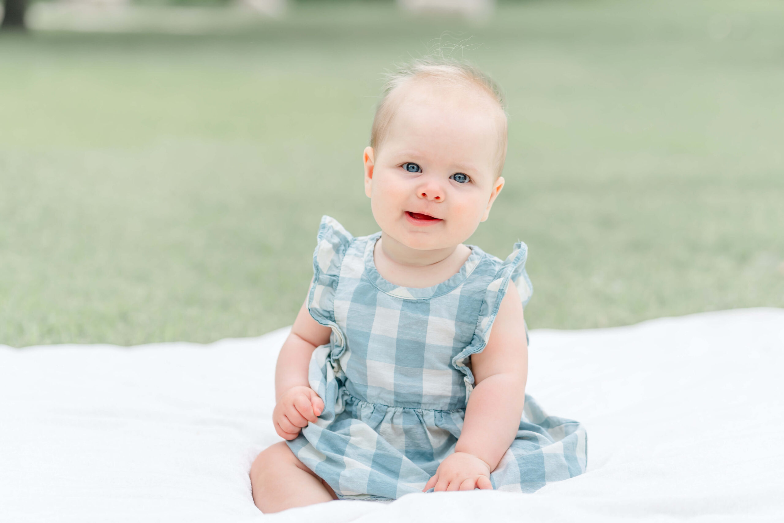 A smiling baby girl in a blue dress sits in a picnic blanket in a park lawn