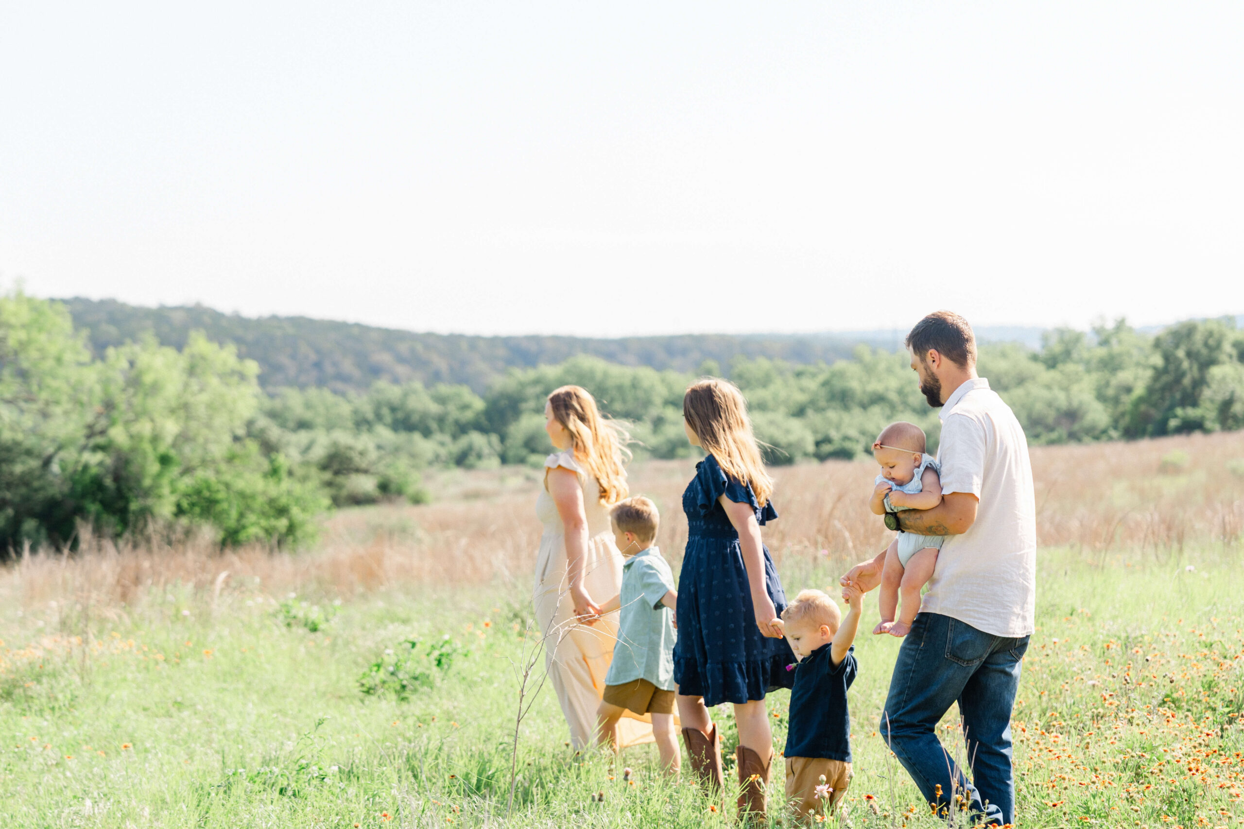 A mom and dad walk in a meadow holding hands with their two toddlers, teen daughter and holding a baby