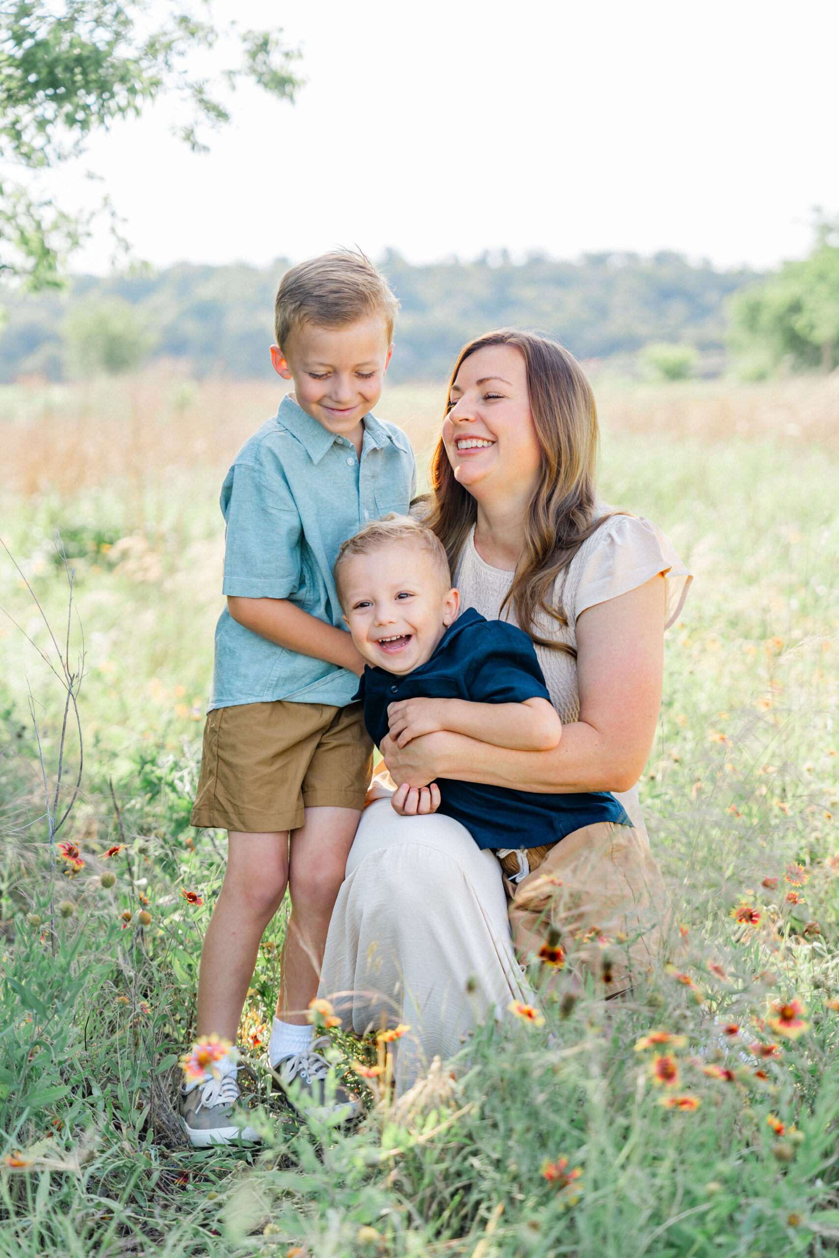 A happy woman kneels in a field of wildflowers laughing with her two toddler sons playing in her lap before visiting toy stores in austin