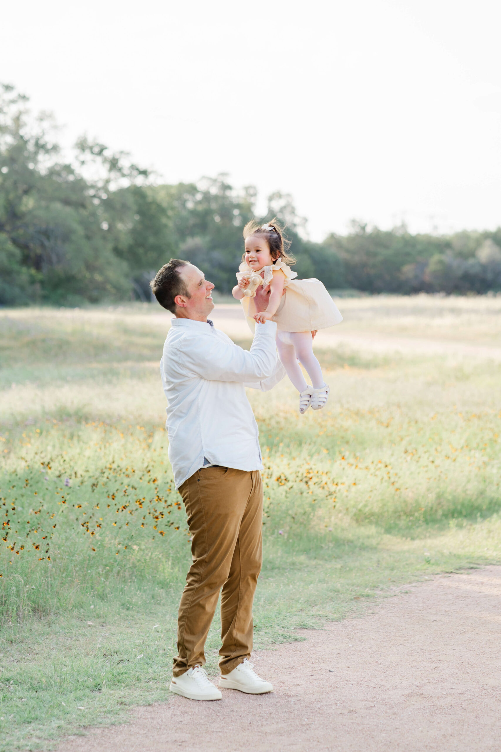 A happy dad in a white shirt lifts his giggling toddler daughter in a yellow dress in a park trail after finding summer camps in austin