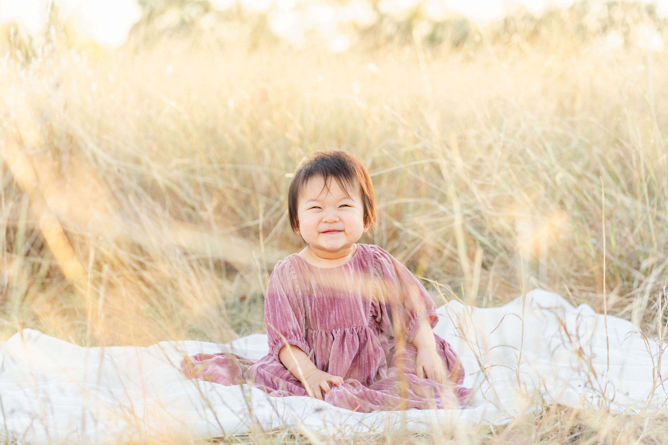 A smiling toddler girl sits on a picnic blanket in a pink dress at sunset