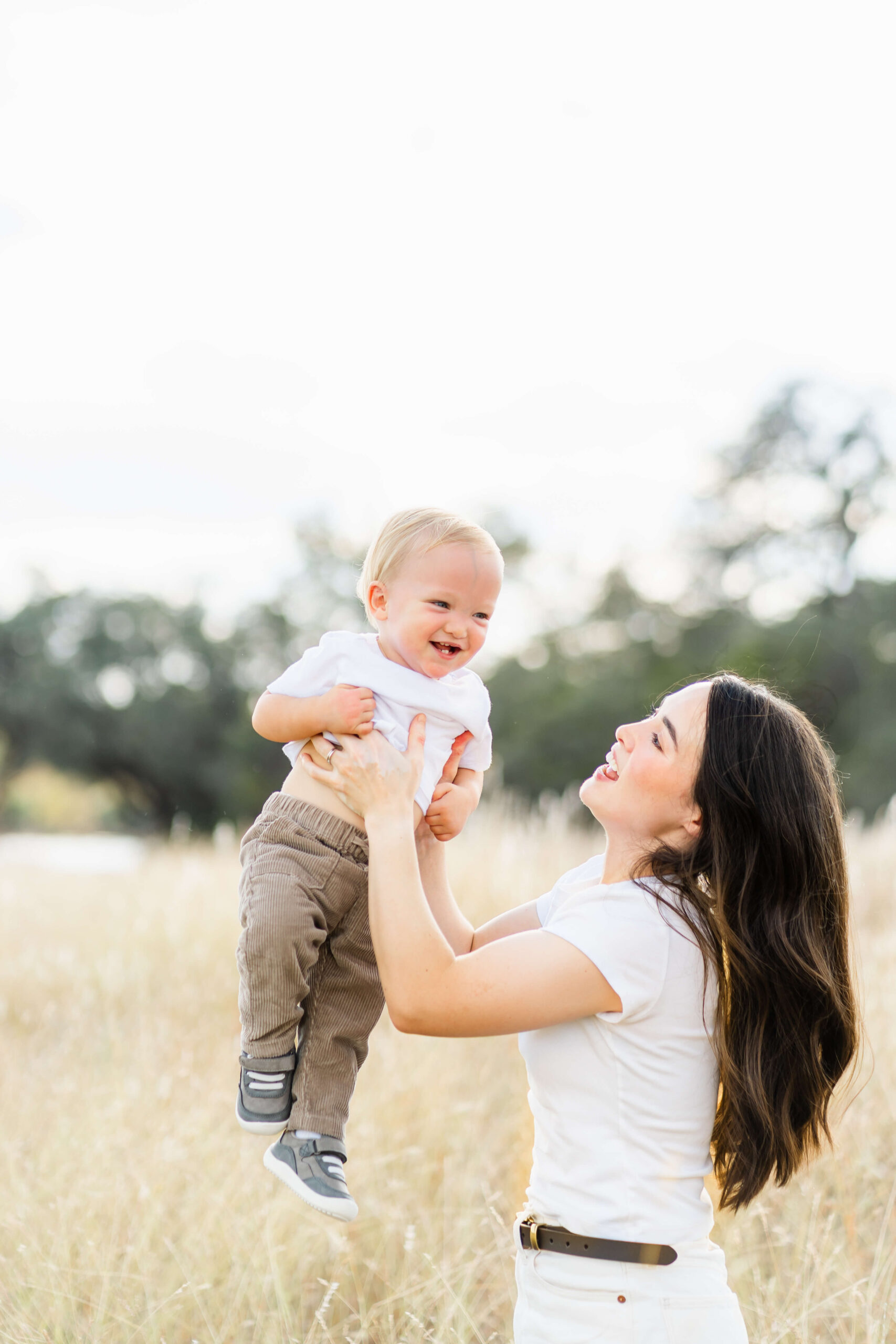 A happy mom in white lifts her giggling toddler in a white shirt while playing in a park