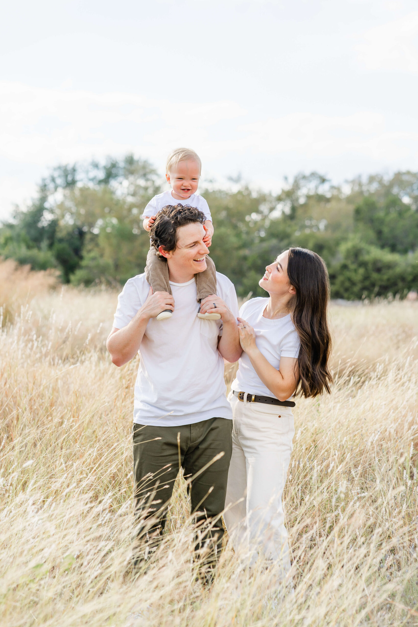 A giggling toddler sits on dad's shoulders on a hike through tall golden grass with mom in white shirts after finding great playgroups in austin
