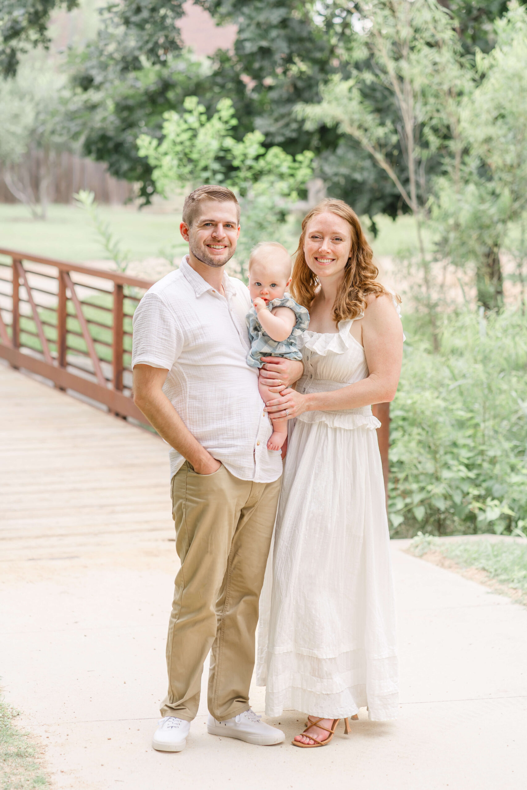 Smiling mom and dad in white and khaki stand in a park by a bridge in a sidewalk with their baby girl in their arms